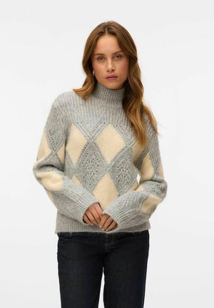 Pull en tricot gris clair avec des motifs en losange crème et des poignets côtelés. Col montant avec des détails en point textured. Associé à un jean foncé.