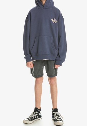 Sudadera de gran tamaño en azul marino con un bolsillo frontal y un detalle gráfico en el pecho, combinada con pantalones cortos grises y zapatillas altas negras.