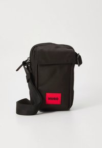 Schwarze rechteckige Umhängetasche mit verstellbarem Riemen und rotem vorderseitigem Aufnäher mit der Aufschrift "HUGO" vor hellem, unifarbenem Hintergrund.