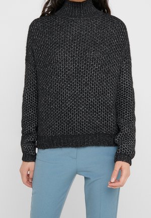 Schwarzer strukturierter Strickpullover mit Rollkragen, kombiniert mit hellblauen, engen Hosen, getragen von einer Person mit langen dunklen Haaren.