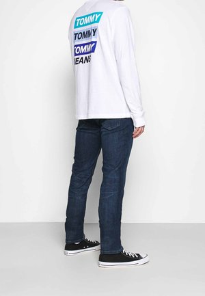 Person trägt ein weißes Langarmshirt mit blauem "Tommy Jeans" Logo auf dem Rücken, dunkelblaue Jeans und schwarze Low-Top-Sneaker und steht seitlich.