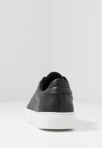 Baskets en cuir noir avec semelle en caoutchouc blanche, vues de dos, sur une surface blanche avec des murs gris unis.