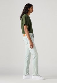 Grønn t-skjorte med korte ermer, lyseblå jeans, stående profil. Myk stoff, vanlig passform, lys vask med lett slitasje, hvite sneakers.