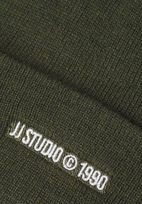 Gebreide groene beanie met ribbelstructuur. Heeft een witte geborduurde logo "JJ STUDIO © 1990" langs de rand.