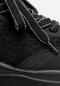 Zapato negro que presenta una combinación de fieltro texturizado y material suave, con cordones negros y con acentos metálicos, además de sutiles detalles de costura.