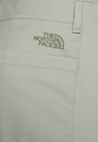 Světlé šedé látky s béžově vyšívaným logem "THE NORTH FACE" a prošitými detaily švů poblíž poutka na opasek.