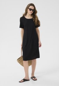 Black short-sleeve dress with a round neckline, two side pockets, and a loose fit. Model carries a woven straw bag and wears slide sandals.