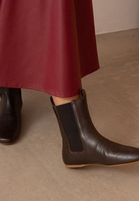 Brązowe skórzane botki na kostkę z elastycznymi panelami po bokach, spiczastymi noskami i płaskimi gumowymi podeszwami, współtworzące zestaw z burgundową spódnicą do kolan.