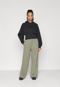 Sieviete ar pītu matu stilu, ģērbusies melnā īsajā kapucē un platgala olīvu zaļās biksēs, stāv pretī vienkāršai gaišai fona.