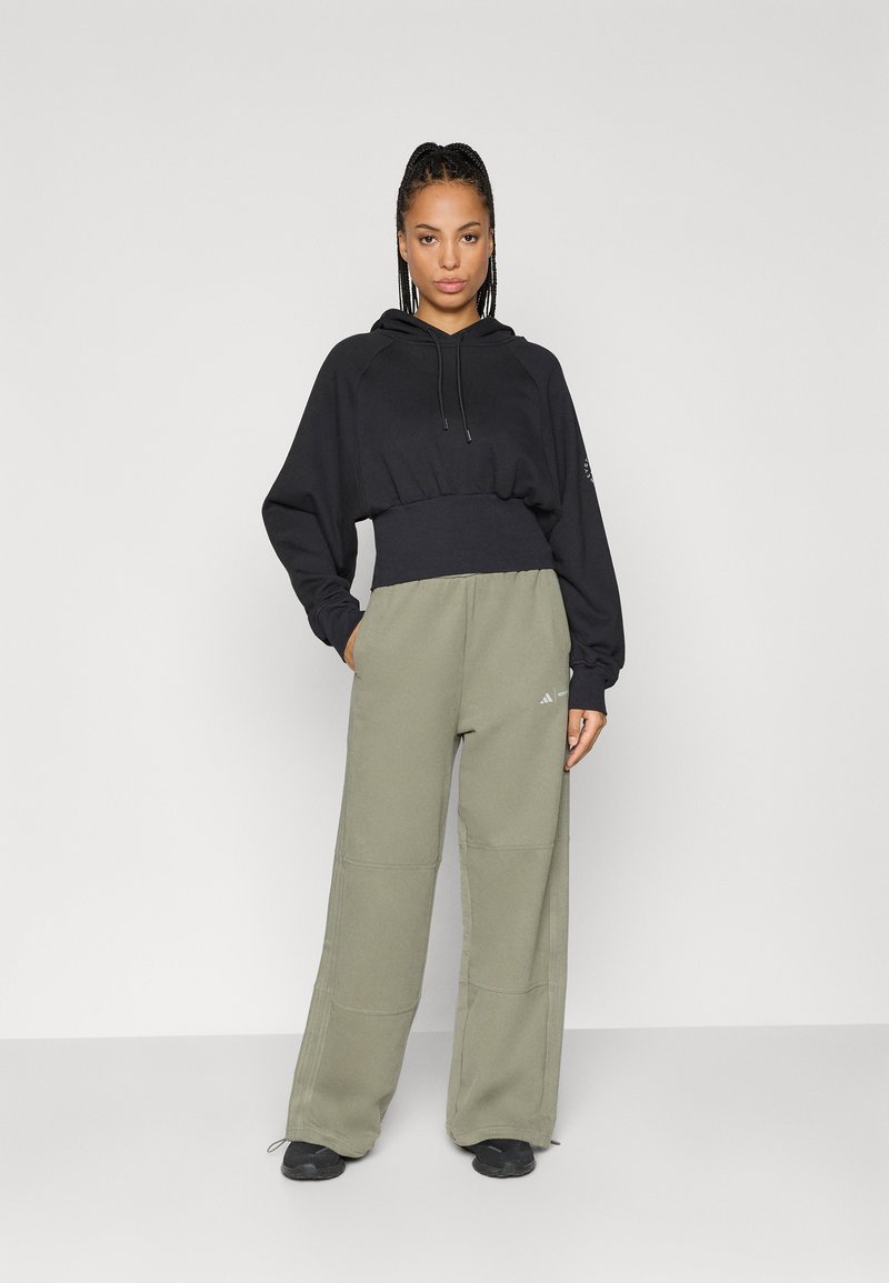 Vrouw met gevlochten haar, gekleed in een zwarte cropped hoodie en olivegroene wijde broek, staande tegen een effen lichte achtergrond.