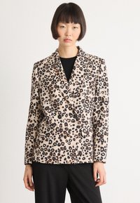 Vrouw met kort, donker haar, gekleed in een beige dierenprint dubbelzijdige blazer over een zwarte top en zwarte broek, staande tegen een witte achtergrond.