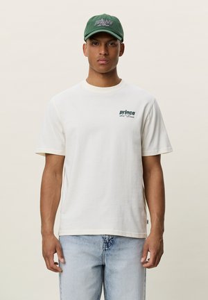 Νέος άνδρας με λευκό t-shirt "prince des Deux", ανοιχτό μπλε τζιν και πράσινο καπέλο "prince des Deux", που στέκεται μπροστά σε απλό φόντο.