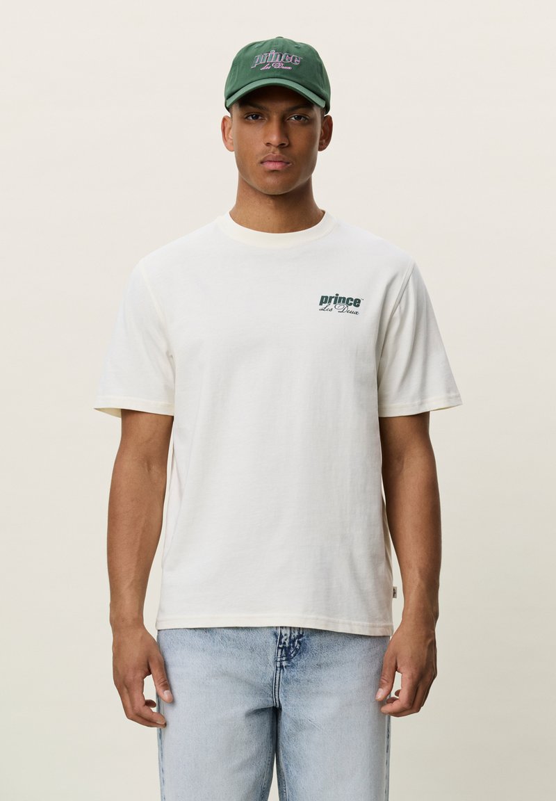 Jeune homme portant un t-shirt blanc « prince des Deux », un jean bleu clair et une casquette verte « prince des Deux », debout devant un fond uni.