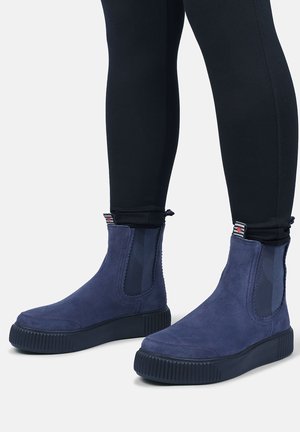 Navy blue suede ankle boots with thick black soles worn with black fitted pants, shown on a neutral light background.
