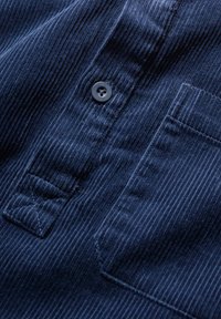 Temno modra kordna tkanina z navpičnimi rebri, opremljena s črnim gumbom in prsno žepno s trikotno zanko.