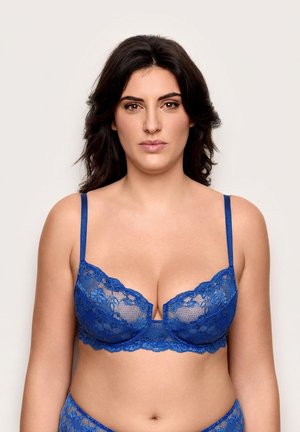 Frau mit dunklen Haaren, die einen blauen Spitzen-BH und passende Unterwäsche vor einem schlichten hellen Hintergrund trägt.