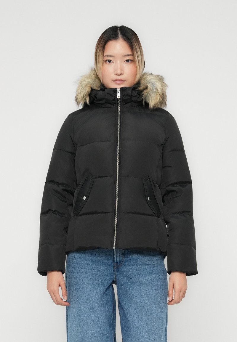 Ung kvinde iført en sort buldredragt med hætte i pels og blå jeans, vendt fremad mod en ensfarvet baggrund.