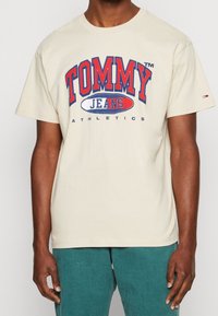 Beige bavlněné tričko se červeným a modrým grafickým motivem "TOMMY JEANS ATHLETICS". Krátké rukávy, kulatý výstřih a malá logo aplikace na rukávu.