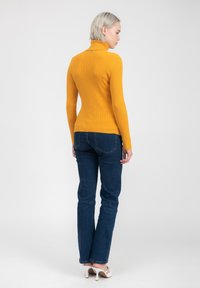 Maglione a collo alto a coste di colore giallo senape con maniche lunghe, abbinato a jeans dritti blu scuro e tacchi bianchi. Tessuto dalla texture liscia.