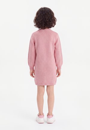 Jeune enfant aux cheveux bouclés portant une robe rose à manches longues et des baskets blanches, debout de dos sur un fond blanc uni.