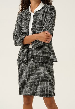 Nainen, jolla on päällään mustavalkoruudullinen tweed-takki ja siihen sopiva hame sekä valkoinen napillinen pusero alla, seisoo käsivarret ristissä.