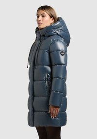 Puffa kabát v tmavě modré barvě s lesklým povrchem, př oversized kapucí, zipovými kapsami a prošívaným vzorem. Logo značky na rukávu.