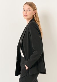 Blazer nero a righe sottili realizzato in tessuto strutturato, con chiusura a un bottone, rever a scoller e tasche frontali.