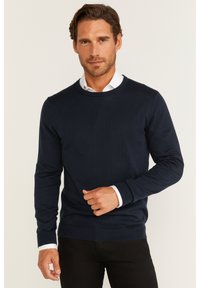 Granatowy sweter okrągły w dekolcie z prążkowanymi mankietami i dołem, z teksturowanym splotem, noszony na białej koszuli z kołnierzykiem.