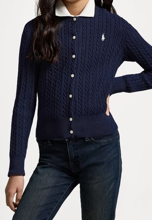 Mujer con un cárdigan de botones de punto con trenza azul marino, con cuello blanco y logotipo bordado en blanco, combinado con jeans oscuros.