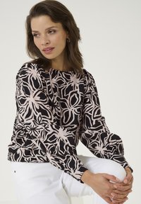 Schwarz-rosafarbene Bluse mit floralem Muster, langen Ärmeln, lockerer Passform, gerippten Bündchen und rundem Halsausschnitt. Kombiniert mit weißen Hosen.