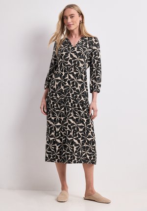Femme portant une robe midi à manches longues noire et beige à motif floral avec des chaussures beiges à enfiler, debout devant un fond clair uni.