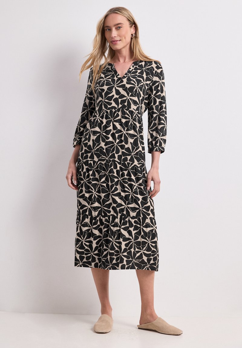 Femme portant une robe midi à manches longues noire et beige à motif floral avec des chaussures beiges à enfiler, debout devant un fond clair uni.