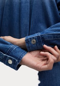 Jasnoniebieskie mankiety dżinsowej kurtki, z zapięciem na guzik z metalowymi elementami. Tkanina ma klasyczną teksturę dżinsu oraz wykończenie szwów.