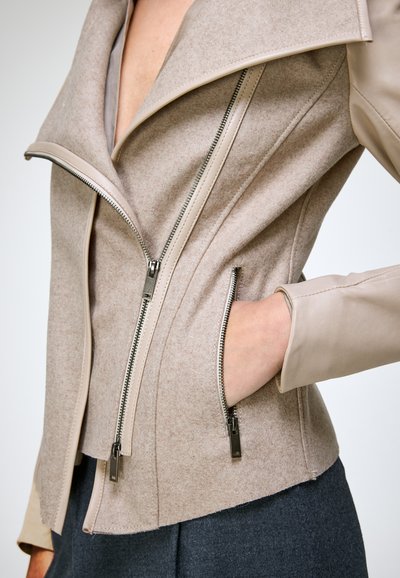 Veste beige en laine et cuir avec fermeture éclair asymétrique et poche zippée, portée sur un pantalon foncé avec la main dans la poche.