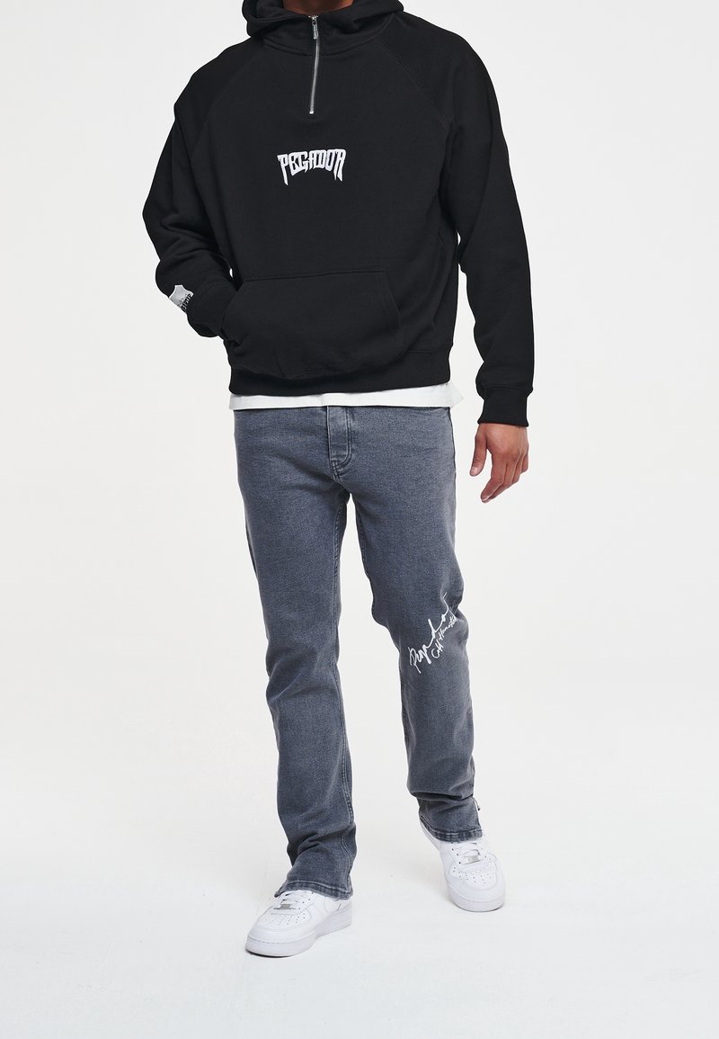 Felpa nera con zip e tasca frontale, logo bianco, abbinata a jeans grigi con dettaglio firmato bianco e sneakers bianche.