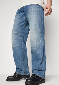 Lockere blaue Jeans aus Denim mit einer leicht verblassten Waschung, die über gebogene Nahtdetails und einen geraden Schnitt verfügt. Kombiniert mit schwarzen Lederschuhen.