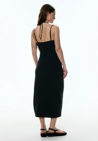 Zwart, aansluitend maxi-jurk met dunne bandjes en een strik op de rug. Gladde stof, enkel-lang met een lichte glans, aangevuld met chunky sandalen.