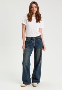 Blauwe wijd uitlopende jeans met vervaagde plekken, vijf zakken en riemlussen, gecombineerd met een eenvoudig wit T-shirt. Gladde stoftextuur.