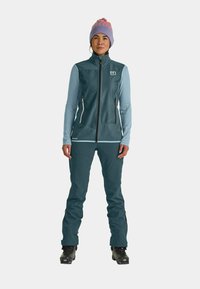 Winteroutfit in Blau und Türkis mit mehreren Schichten, bestehend aus einer Reißverschlussweste und einer Hose; verfügt über Reißverschlusstaschen, strukturierten Stoff und eine gemusterte lila Mütze.