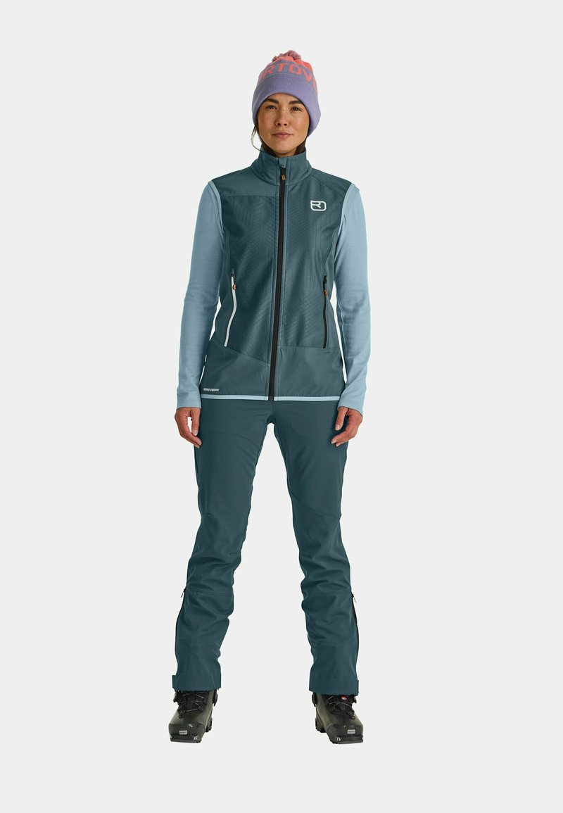 Winteroutfit in Blau und Türkis mit mehreren Schichten, bestehend aus einer Reißverschlussweste und einer Hose; verfügt über Reißverschlusstaschen, strukturierten Stoff und eine gemusterte lila Mütze.