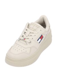 Krémově zbarvená teniska s perforovanou špičkou, zavazováním na tkaničky, texturovanými stranami a výrazným logem s červenými, bílými a modrými vodorovnými pruhy.