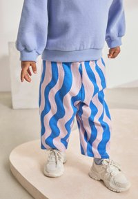 Blaue und rosa gestreifte Hosen mit wellenförmigen Mustern, kombiniert mit einem hellblauen Sweatshirt. Weiße Sneakers runden das Outfit ab.