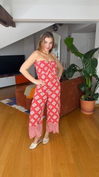 Γυναίκα με κόκκινη και λευκή ολόσωμη φόρμα χωρίς τιράντες με σχέδιο και λευκές παντόφλες στέκεται με τα χέρια στη μέση σε ένα σαλόνι με φυτά και έπιπλα.