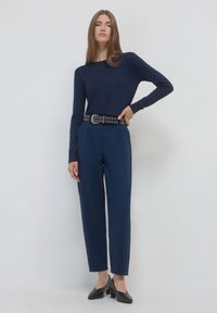 Giovane donna che indossa una maglietta a maniche lunghe blu navy, pantaloni blu navy a vita alta con una cintura nera e scarpe nere con tacco basso, in piedi contro un muro bianco.