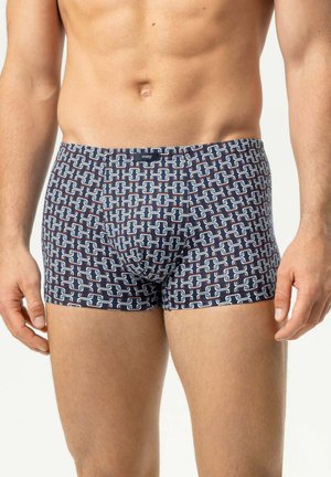 Männlicher Oberkörper, der marineblaue Boxershorts mit weißem und rotem geometrischem Kettenmuster trägt, steht mit locker an den Seiten hängenden Armen.