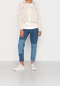 Chaqueta de corduroy beis con dos bolsillos en el pecho, camiseta blanca con estampado, jeans de mezclilla azul con patchwork y zapatillas beis con cordones. Conjunto informal.