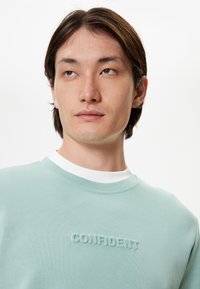 Sudadera verde menta con cuello redondo acanalado y sutil texto en relieve "CONFIDENT" en el pecho; textura suave y corte relajado.