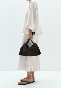 Grand sac à main en suède marron avec une forme triangulaire large et des double poignées supérieures, tenu par une personne portant une tenue blanche ample et des sandales.