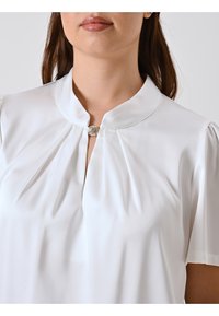 Blusa bianca in tessuto liscio, con colletto rialzato, parte anteriore plissè e una chiusura decorativa al collo. Maniche corte.