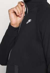 Ruka muže, která stahuje zip černé bundy Nike s bílým logem na hrudi a viditelnými švy.